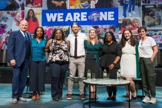 A group poses together for a group shot in front of a sign that says "We Are One"