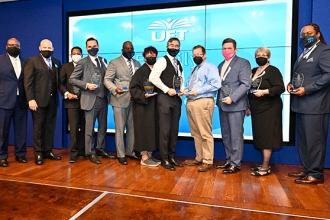 11 people, all masked pose for a group photo at the stage. 9 of the people have awards in their hands. 