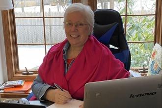 A woman with white hair and a pink shirt smiles while sitting at a desk.