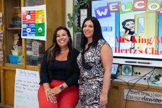 Two teachers stand in front of a SmartBoard that reads "Welcome to Mrs. King/Ms. Hertz's class"