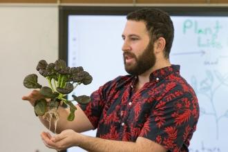 A white man with a beard wearing a flower-patterned shirt holds a leafy plant