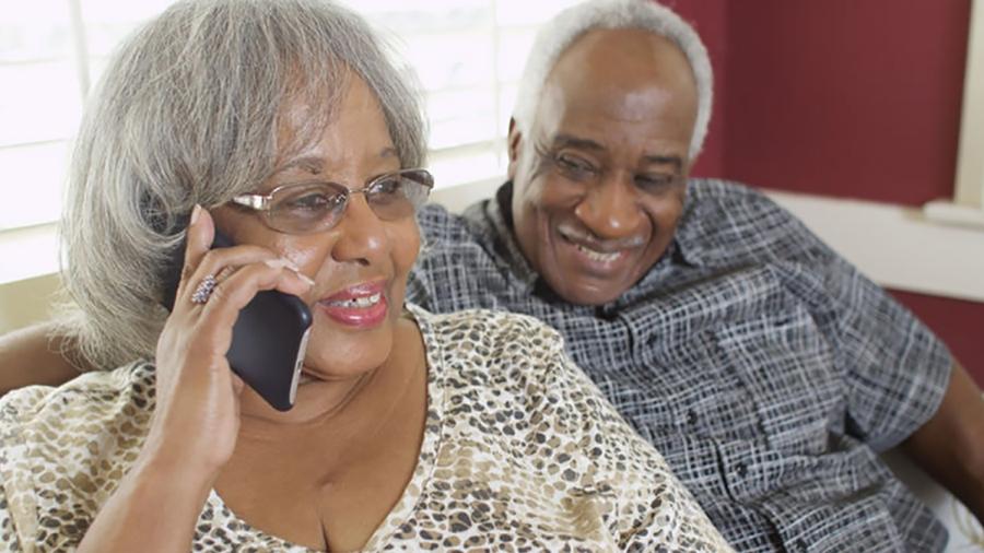 Elderly man and woman sitting next to each other smiling while woman speaks on the phone
