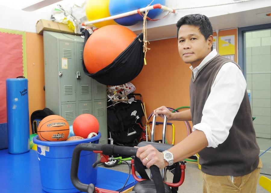A man stands with an adaptive exercise device in a room full of exercise balls and gym equipment