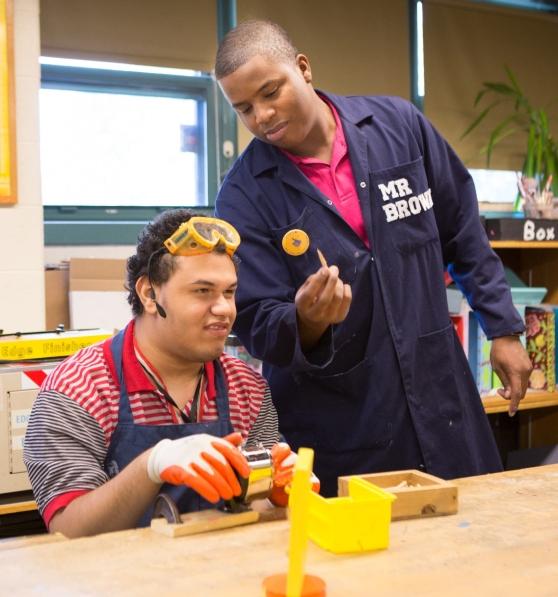 an educator in a jumpsuit that says 'Mr. Brown' assists an adolescent student wearing safety goggles in a wood shop classroom