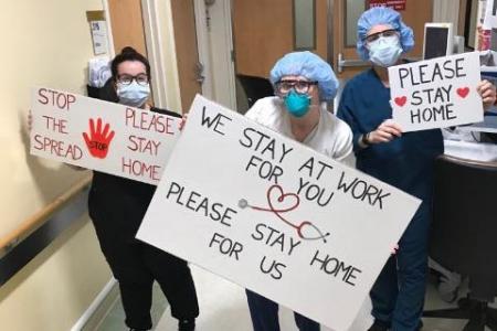 Nurses wearing masks holding signs that say "We stay at work for you, stay home for us"