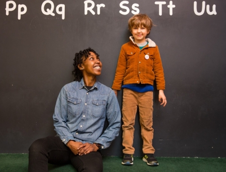 adult sits next to child in front of a blackboard with the ABCs displayed. adult is smiling at child, who is standing.