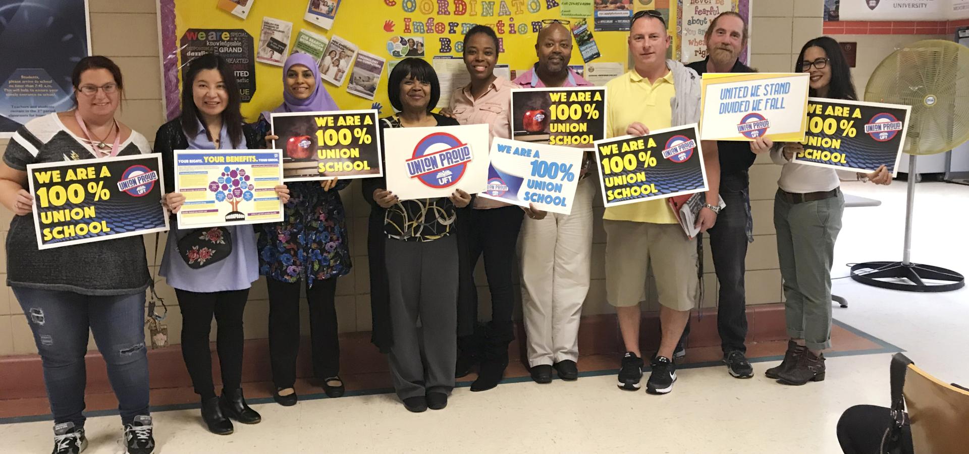 Group of diverse educators holding UFT signs that say "We are a 100% union school" and "Union Proud"