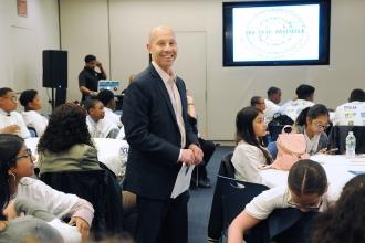 Man standing in a room with children seated, smiles at the camera
