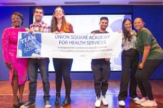 A group of members on stage hold up a banner for their school