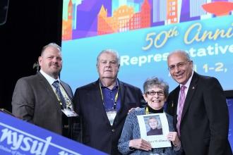 3 older men and an older woman pose for a photo while the woman holds a piece of paper with her name on it 