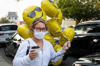 A woman wearing a white mask and white zip-hoodie walks down the street with coffee in one hand and balloons with smiley faces wearing sunglasses. 