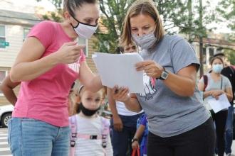Two teachers both wearing masks look down at a piece of paper with a list of students. The teacher on the left is wearing a pink Puma shirt and a pink and black mask while the teacher next to her is wearing a grey t shirt and mask. 