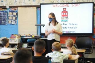 A teacher wearing a blue camo mask is in her class and interacting with students also wearing masks. 