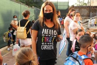 A teacher wearing a shirt reading "Hello First Grade" is centered in the frame and posing while wearing an all black mask. 