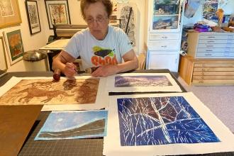 An elderly woman sits by a table in her art studio surrounded by her drawings and paintings.