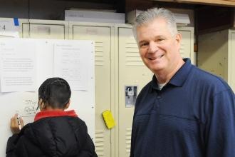 A man smiles for the camera while students in the background work on a whiteboard 