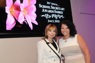 2 women pose for a photo in front of a television screen reading "36th annual School Secretary Awards Soiree"