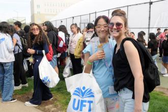 Two women with glasses pose for a photo amongst a crowd of many others in a field with a fence around it, one holding a UFT swag bag