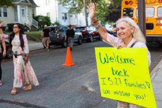 A woman holds a sign reading "Welcome back, IS 27 families, we missed you!"