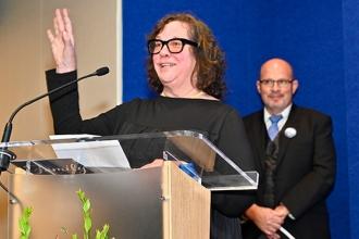 A woman with glasses is speaking at a podium while raising her hand, a man watches in the background. 