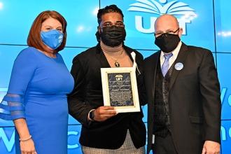 Three people pose for a group photo, all masked. The man in the middle is holding an award. 