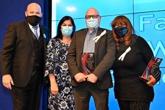 Four people pose for a group photo, all masked. The two people on the right are holding awards. 