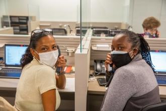 Two women both holding phones and wearing glasses on top of their heads smile behind masks. 