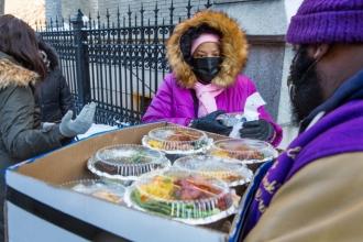 A woman looks to assist a man holding a box of food. 