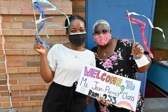 Two women with short hair are wearing masks and waving their hands. They are also holding a sign reading "Welcome to Ms. Jean-Pierre's Class" in rainbow lettering 