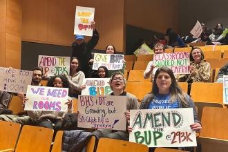 Members sitting in an auditorium hold up signs demanding reallocation of charter school space. 