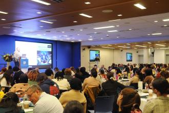 A general crowd photo of Shanker Hall showing some of the 1300+ attendees as someone on the stage (left side), gives a presentation. 