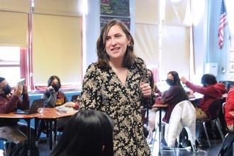 Female teacher standing in room with others who are sitting.