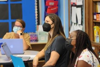 Image shows three teachers seated in a classroom, wearing masks and looking toward a screen