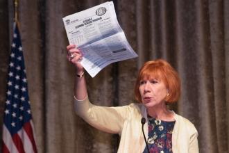 Mature red headed woman stands at podium holding a newspaper in hand
