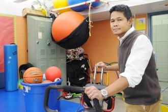 A man stands with an adaptive exercise device in a room full of exercise balls and gym equipment