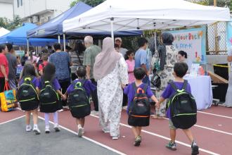 Young students — all holding hands and wearing matching shirts and their new backpacks — are guided through the 5th annual UFT Staten Island Back-to-School Barbecue. 
