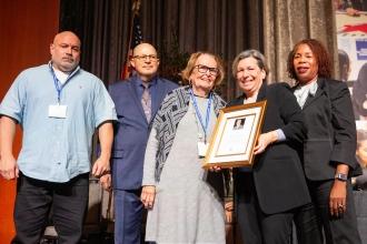 Christopher Jones (far left), son of the late former UFT Vice President for Middle Schools Ron Jones, and Jones’ widow, UFT retiree Frieda Jones (middle), accept an In Memoriam plaque for Jones, who died in May 2025, from Mulgrew, AFT President Randi Weingarten (second from right) and UFT Vice President for Elementary Schools Karen Alford.