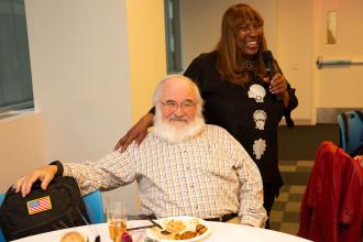 A woman stands next to a man sitting at a table