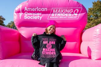 A teacher holds up a breast cancer awareness t-shirt while sitting on a large inflatable chair. 