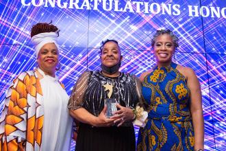 Three women pose for a photo while on stage. The person in the middle is holding a plaque 