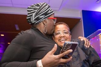 A woman on the left embraces the woman on the right with a kiss to the top of her head