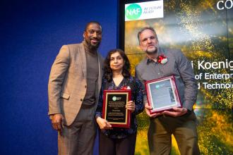 A man on the left poses with two award winners to his right on the stage.