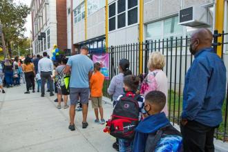 Groups of parents and students are lined up waiting for the school doors to open.
