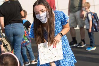 A teacher in a blue floral dress and white mask bends down to introduce herself to a student.