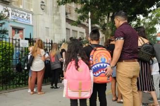 A group of parents and students waiting for the school to open. Two students are wearing backpacks, one is pink with stripes and the other is an orange shark. 