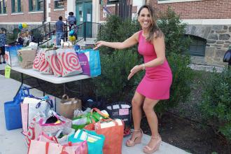 A teacher wearing a pink dress displays a table full of school supplies
