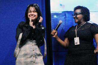 A woman on the left claps as the woman on the right hands her a microphone