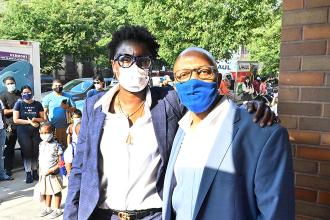 Two teachers both wearing navy suits and glasses pose for a picture in front of a school building.