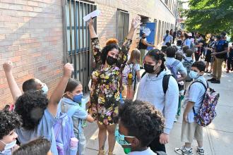 A teacher wearing a floral dress is surrounded by students as she raises her arms in joy. 