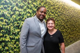 A man and woman pose for a photo against a wall painted with leaves.